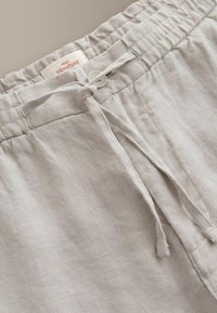 Pantalon en lin beige clair avec ceinture élastique, lien de serrage et fermeture par un seul bouton blanc, étiquette visible à l'intérieur de la ceinture.