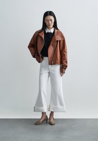 Casaco bomber de couro castanho com gola oversize, suéter preto, jeans brancos de perna larga com bainhas desfiadas e sapatos de salto alto castanhos.