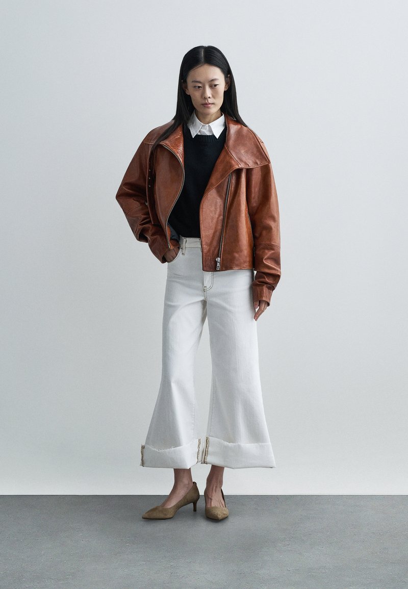 Casaco bomber de couro castanho com gola oversize, suéter preto, jeans brancos de perna larga com bainhas desfiadas e sapatos de salto alto castanhos.