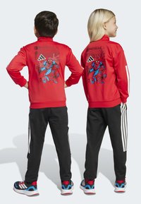 Rode trainingsjack met spinnenafbeelding op de achterkant, zwarte broek met witte strepen en kleurrijke schoenen, die een sportieve, casual uitstraling laten zien.