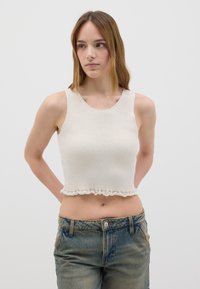 Top corto a coste color crema con orlo ondulato, abbinato a jeans blu sbiaditi. Il top presenta spalle larghe e scollatura rotonda.