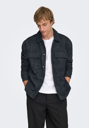 Temno siva jakna iz umetnega žameta z gumbi, z dvema žepoma na prsih, ovratnikom in dolgimi rokavi. Nositvena preko bele majice, kombinirana s črnimi hlačami.