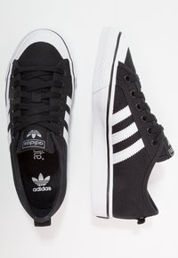 Black canvas sneakers with white rubber sole, featuring three white stripes, black laces, and a textured toe cap. Logo on insole.