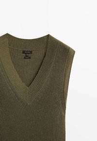 Gilet tricoté sans manches vert olive avec col en V profond et col et emmanchures côtelés, posé à plat sur fond blanc.