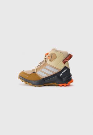 Botte de randonnée beige avec des accents gris, des bandes réfléchissantes et des accessoires orange. Elle possède une semelle robuste et un soutien de la cheville. Conçue pour une utilisation en extérieur.