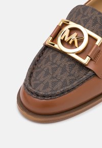 Close-up van een bruine leren instapper met een bovenkant van getextureerde stof, die een herhalend logo-patroon en een gouden, ronde gesp-detail heeft.