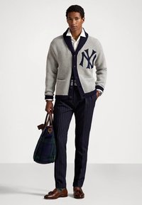 Man wearing gray cardigan with navy trim and large NY logo, white shirt, navy pinstripe pants, brown loafers, holding dark plaid tote bag.