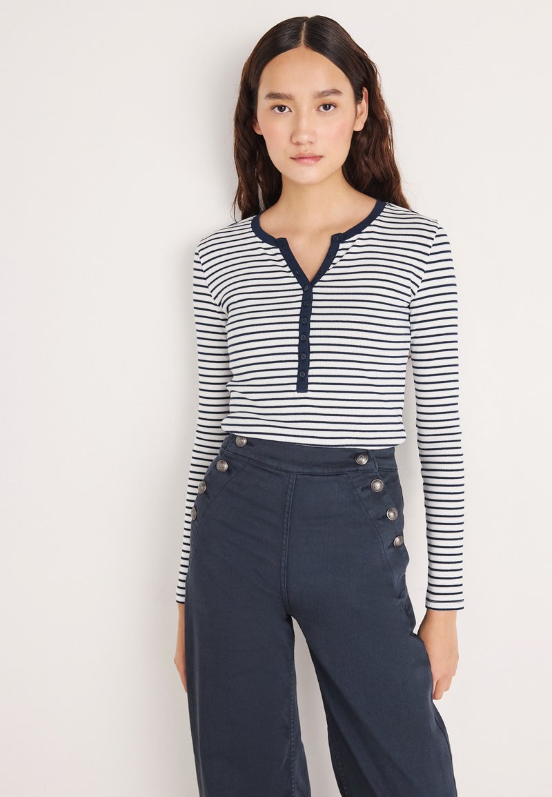 Μακρυμάνικο, ριγέ τοπ σε λευκό και ναυτικό μπλε με λαιμόκοψη henley με κουμπιά. Συνδυάζεται με παντελόνια ψηλής μέσης σε ναυτικό μπλε με ασημένια κουμπιά.