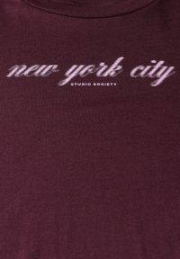Bordová trička s hladkou texturou a bílým nápisem "new york city" s gradientním efektem, pod nímž se nachází "STUDIO SOCIETY".