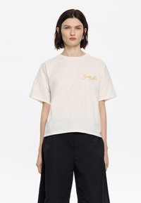 Crème-kleurig katoenen t-shirt met korte mouwen en een relaxte pasvorm, met gouden geborduurde tekst op de linkerborst.