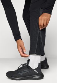 Pantalons de sport noirs avec des zips aux chevilles, associés à des chaussures noires avec une surface texturée et le logo "Terrex".