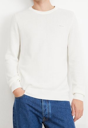 Mężczyzna w białym, teksturowanym swetrze z długim rękawem i okrągłym dekoltem oraz niebieskich dżinsach, jedna ręka w kieszeni, na jednolitym tle.