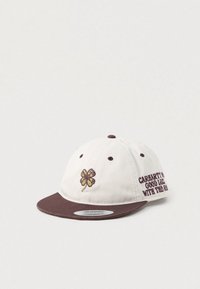 Casquette snapback blanche et brune avec un trèfle à quatre feuilles brodé et le texte "Carhartt WIP Bonne chance avec la course" sur le panneau latéral.
