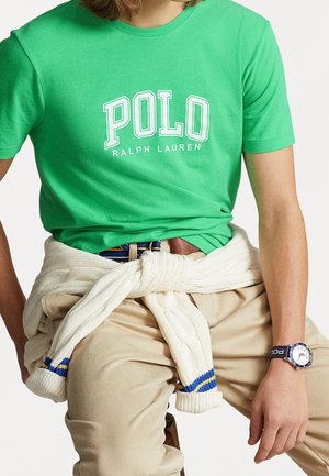 Zielona bawełniana koszulka z nadrukiem "POLO RALPH LAUREN" w białym kolorze. Kremowy sweter związany w talii. Jasnobeżowe spodnie. Niebieski zegarek na nadgarstku.