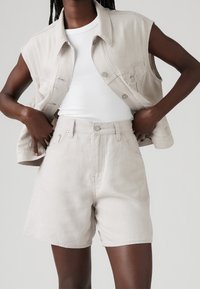 Personne porte un short en denim beige clair, un débardeur blanc côtelé et une veste sans manches en denim assortie, debout, les mains sur les hanches.