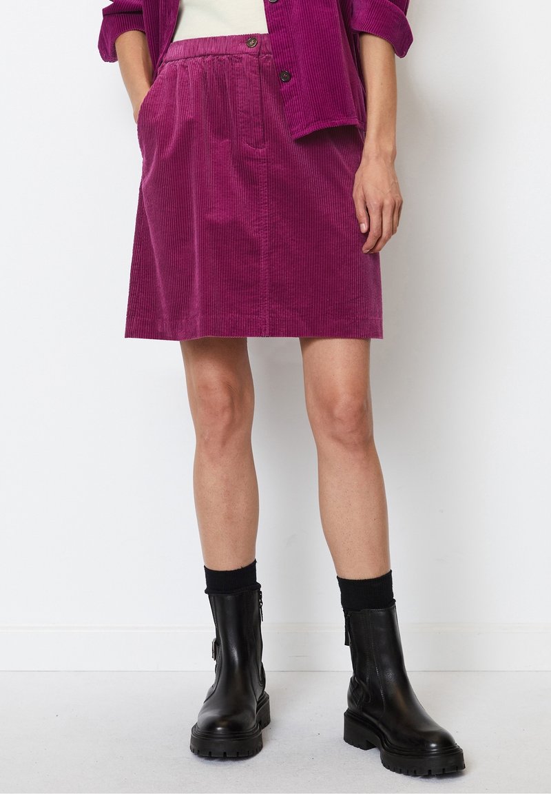 Jupe en velours côtelé violet avec un devant plissé, des poches latérales et une taille avec un fermoir à bouton. Associée à des bottes noires et des chaussettes.