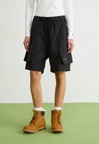 Schwarze Cargo-Shorts mit elastischem Bund, verstellbarem Kordelzug und Seitentaschen. Kombiniert mit braunen Schnürstiefeln und weißen Kniestrümpfen.