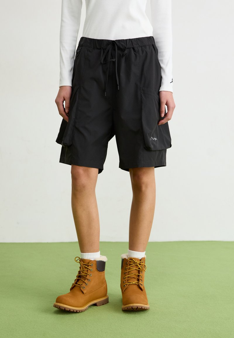 Schwarze Cargo-Shorts mit elastischem Bund, verstellbarem Kordelzug und Seitentaschen. Kombiniert mit braunen Schnürstiefeln und weißen Kniestrümpfen.