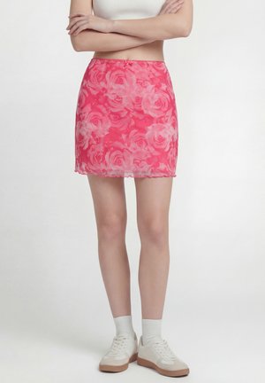 Kvinde iført en lyserød blomsterprint mini-nederdel, hvid crop top, hvide sokker og beige sneakers, står med armene over kors.