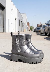 Bottines matelassées noires à semelles épaisses à crampons et fermeture éclair avec logo Buffalo, placées sur un trottoir urbain avec des bâtiments et des voitures garées en arrière-plan.