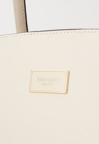 Cream leather tote bag featuring a gold Kate Spade logo plate. The smooth surface has a fine texture, with visible stitching along the edges.