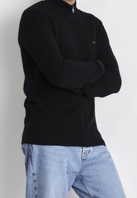 Schwarzer Reißverschluss-Pullover mit hohem Kragen, strukturiertem Strickstoff, enger Passform und gerippten Bündchen, kombiniert mit hellblauen Jeans.