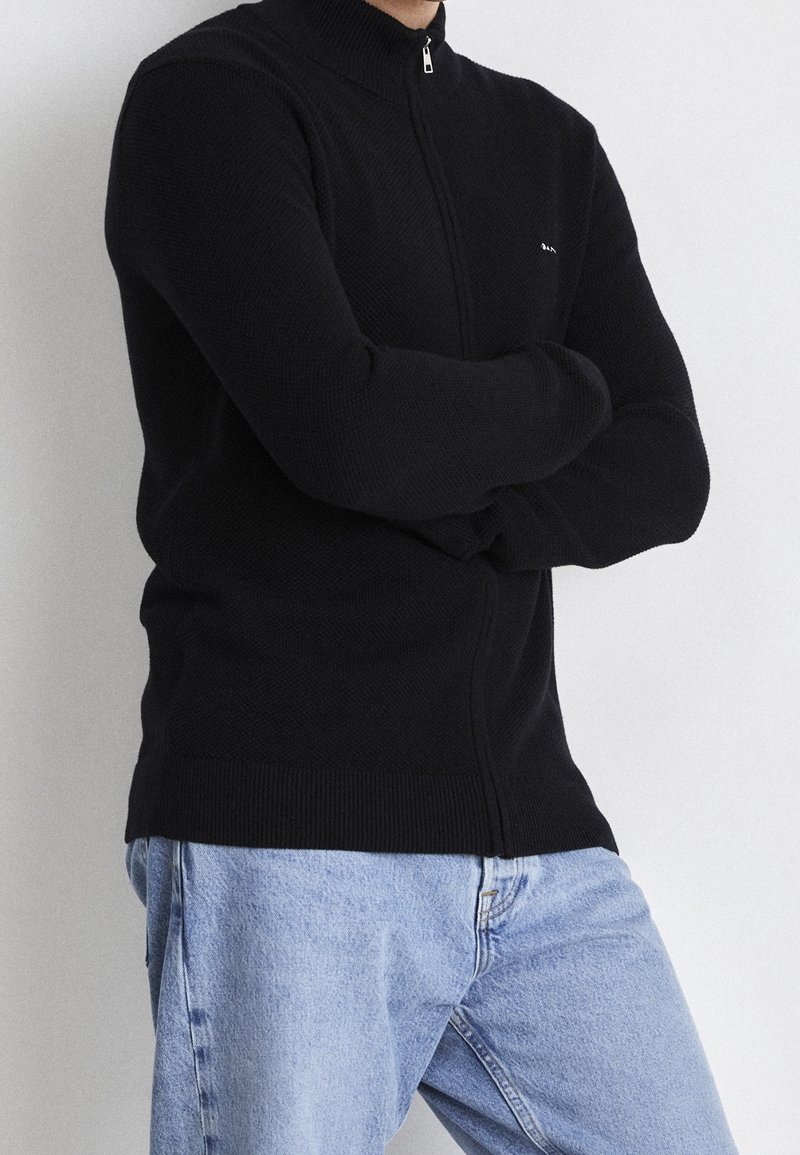 Schwarzer Reißverschluss-Pullover mit hohem Kragen, strukturiertem Strickstoff, enger Passform und gerippten Bündchen, kombiniert mit hellblauen Jeans.