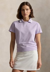 Ženska, oblečena v svetlo vijolično polo majico s kratkimi rokavi, belimi črtami na ovratniku in belo krilo, stoji pred nevtralnim ozadjem.