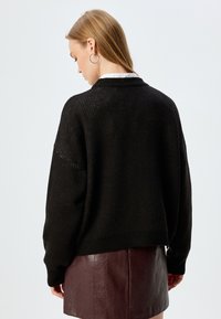 Zwarte gebreide trui met een losse pasvorm, geribbelde zoom en open breistekstuur. Heeft een kraag en valt op de taille. Gepaard met een bordeaux leren rok.