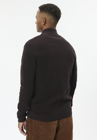 Man with short curly hair wearing a dark brown long-sleeve sweater and brown corduroy pants, facing away against a white background.