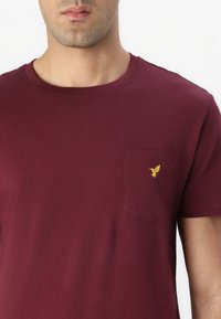 Burgundy short-sleeve t-shirt in cotton with a chest pocket featuring a small yellow bird embroidery detail. Smooth texture and classic fit.