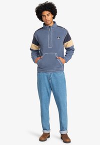 Polarowa bluza w kolorze niebieskim z beżowymi i granatowymi akcentami, zamek błyskawiczny do połowy, przednia kieszeń i teksturowany materiał. Noszona z jasnymi dżinsami i brązowymi butami.