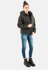 Sort pufferjakke med hætte, lynlåslukning og tekstureret stof. Kombineret med tætsiddende blå jeans og sorte snøreboldede støvler.