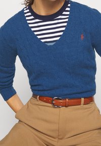 Blue cable-knit sweater with V-neck, worn over a navy and white striped T-shirt. Brown belt with silver buckle over tan high-waisted pants.