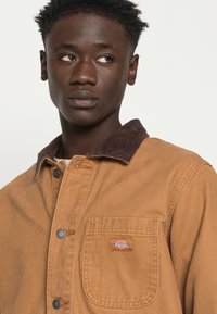 Braune Canvas-Jacke mit einem Cordkragen, ausgestattet mit einer Brusttasche und einem Logo-Patch. Strukturierter Stoff und metallene Druckknöpfe sind sichtbar.