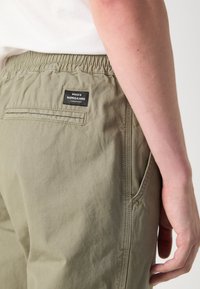 Olivgrüne Jogginghose mit elastischem Bund, Seitentaschen und einer Gesäßtasche mit einem schwarzen Etikett mit weißem Text.