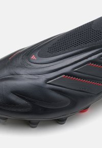 Schwarzer Fußballschuh mit strukturierter synthetischer Oberseite, versehen mit roten Akzenten, einem Mesh-Bereich und Stollen an der Sohle für Grip.