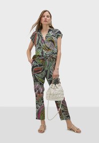 Two-piece outfit featuring a short-sleeve button-up shirt and tailored pants in a dark green fabric with colorful abstract leaf patterns. Complemented by a textured white handbag.
