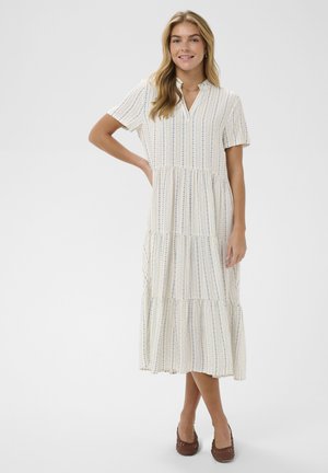 Nő áll, egyik kezét a csípőjén tartva, fehér, kék függőleges csíkozású midi ruhát viselve, barna szövött lapos cipőben, egyszerű háttér előtt.