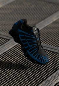 Black and blue athletic sneaker with thick, segmented sole resting on a metal grated surface, highlighting shoe details and design.