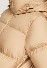 Beige pufferjack met een capuchon; voorzien van gequiltte vulling, trekkoordaccenten en een soepele, lichte textuur. Zichtbare ritssluiting.