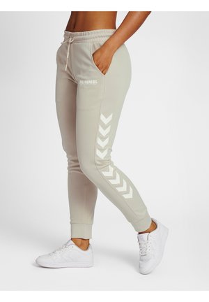 Vue latérale d'une personne portant un jogging beige Hummel avec des bandes chevron blanches et des baskets blanches, debout avec une main dans la poche.