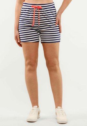 Osoba ubrana w granatowo-białe szorty w paski z czerwonym sznurkiem i białe trampki, stojąca na jednolitym tle.