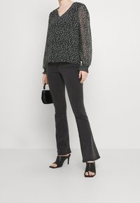 Svart blommig blus med långa ärmar och V-ringning, kombinerad med grå vidbyxor. Modellen bär svarta öppna klackar och håller en liten svart väska.