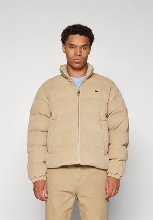 Chaqueta puff de pana beige con cuello alto, cierre de cremallera y un sutil logo de la marca. Presenta puños acanalados y una textura suave.