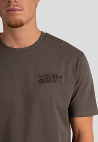 Camiseta de algodón marrón con cuello redondo y mangas cortas que presenta un logotipo bordado en negro "WRONG FRIENDS" en el pecho.