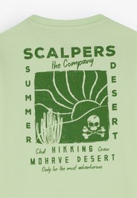 Lichtgroene T-shirt met groene grafische tekst. Het ontwerp omvat illustraties van een woestijnlandschap, een schedel en cactussen met de branding "SCALPERS".