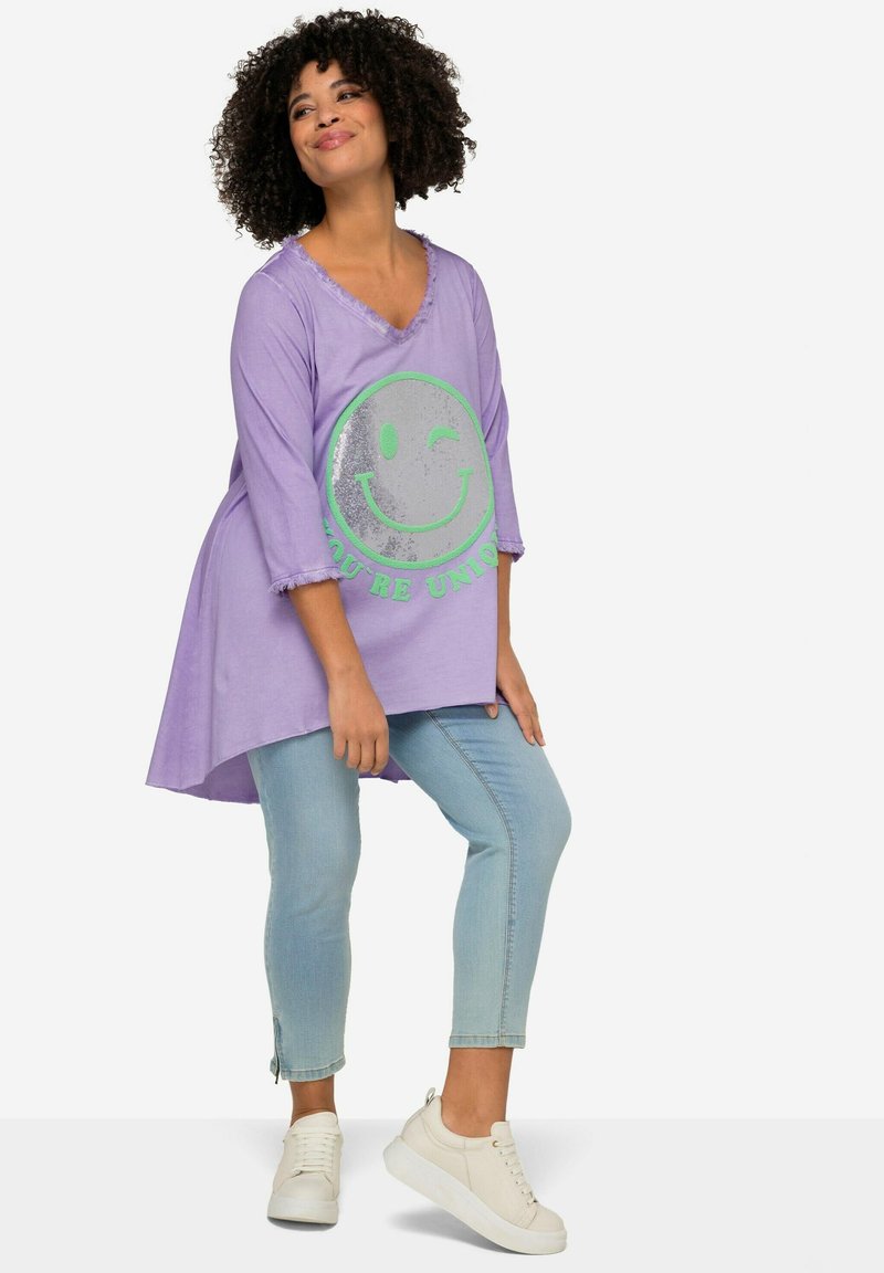 Paarse oversized top met V-hals en gefranjerde mouwen, heeft een zilveren smiley ontwerp met groene tekst op de voorkant. Gecombineerd met lichtblauwe jeans en witte sneakers.