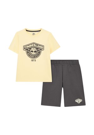 T-shirt Timberland jaune à manches courtes avec logo et texte "1973", associé à un short gris foncé à taille élastique avec un petit logo Timberland.