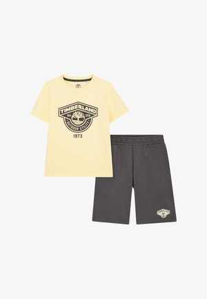 Maglietta gialla Timberland a maniche corte con logo e scritta "1973", abbinata a pantaloncini grigio scuro con vita elastica e piccolo logo Timberland.